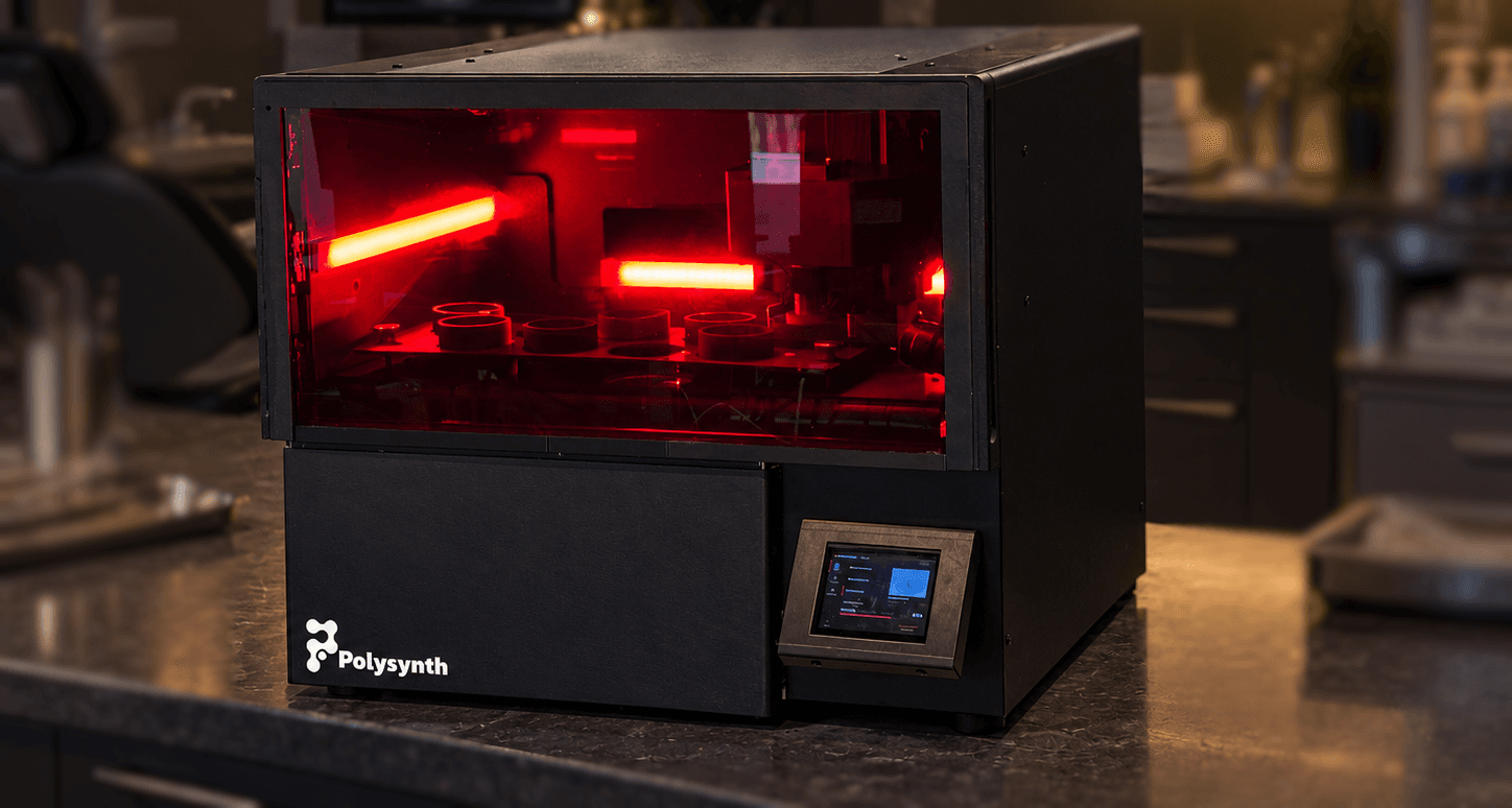 The Polysynth multi-material printer on a dental clinic counter, its red-lit build chamber holding several resin vats.