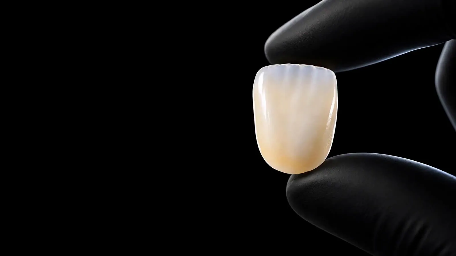 A gloved hand holding a layered zirconia crown, showing a translucent enamel layer over warmer dentin tones.